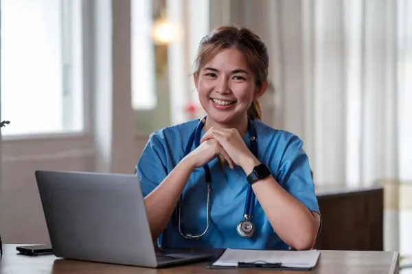 A smiling woman in scrubs sits at a desk with a laptop