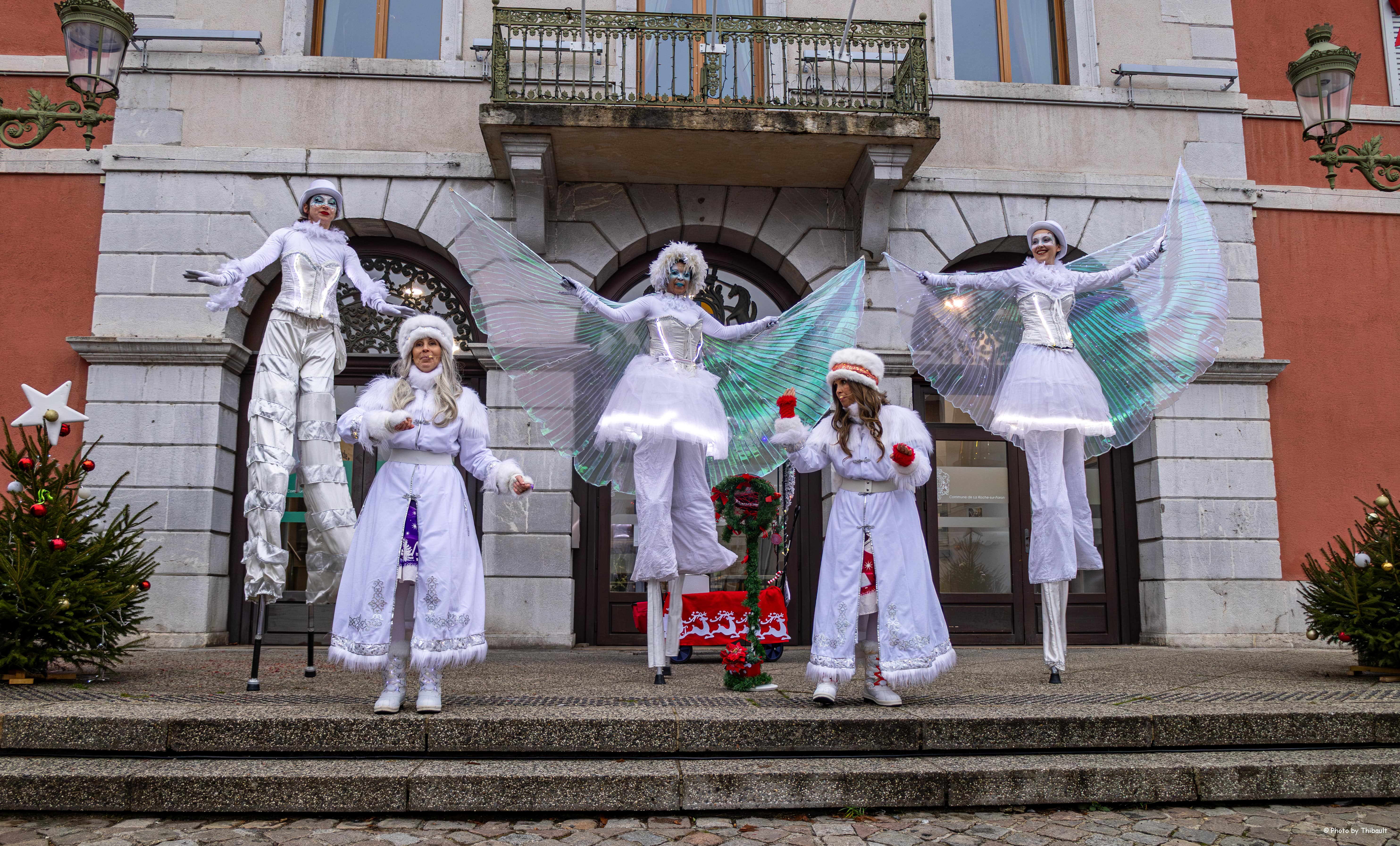 Cinq artistes en costumes blancs d'anges et manteaux d'hiver avec des ailes lumineuses, se tiennent devant un bâtiment en pierre décoré pour Noël.