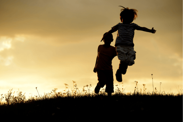 Niños a contraluz saltando en un césped con el fondo del atardecer detrás.