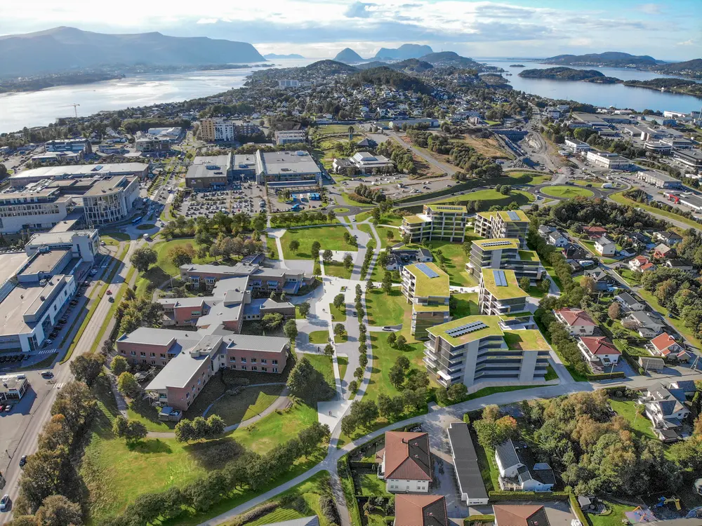 Flyfoto av en Moa Hageby med moderne bygninger, grøntområder og parkeringsplasser, omgitt av vann, fjell og øyer i det fjerne, under en delvis overskyet himmel