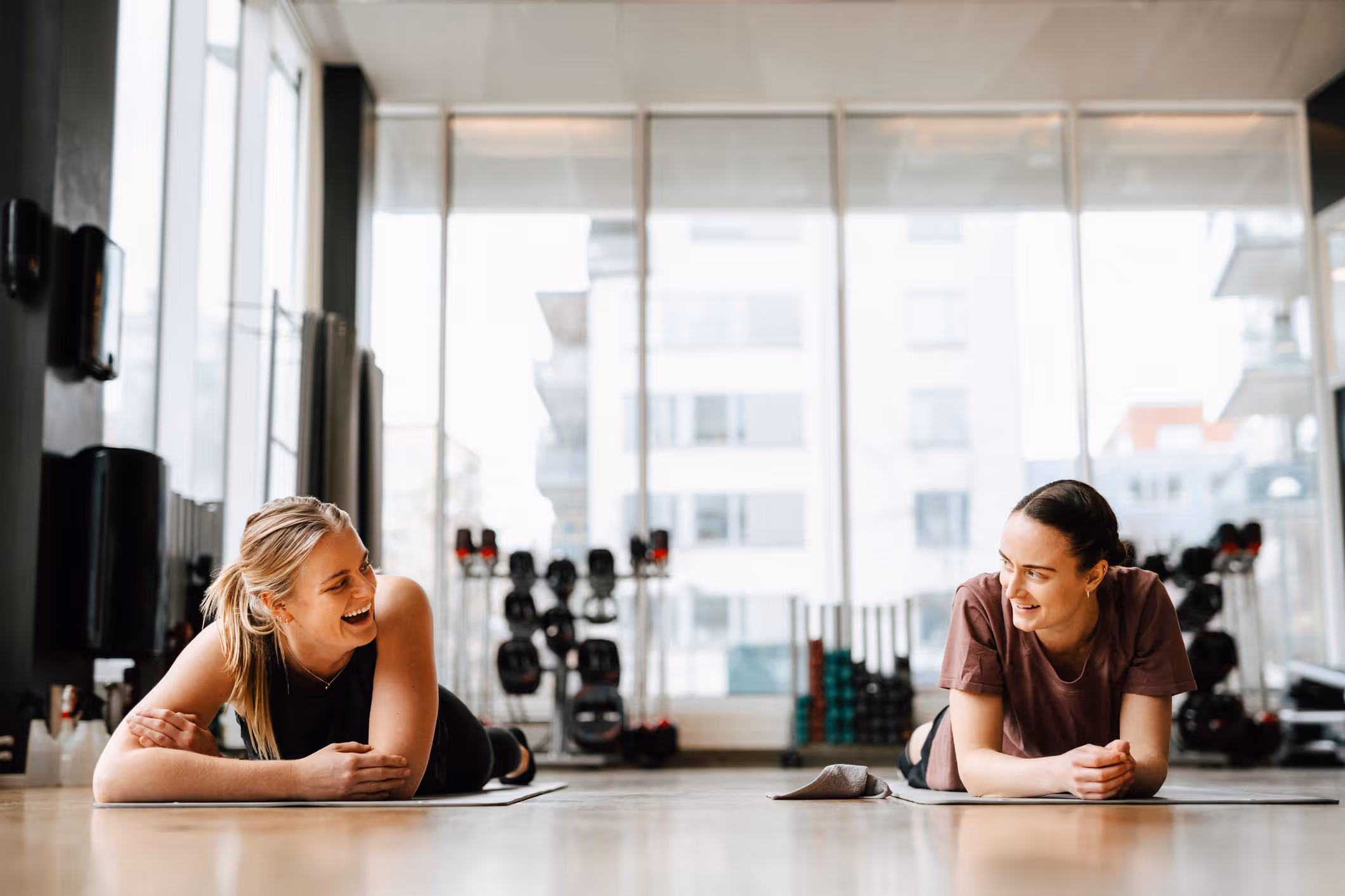 To kvinner ligger på yogamatter i et treningsstudio, mens de smiler og snakker med hverandre. I bakgrunnen ser man treningsutstyr og store vinduer med bygninger utenfor.