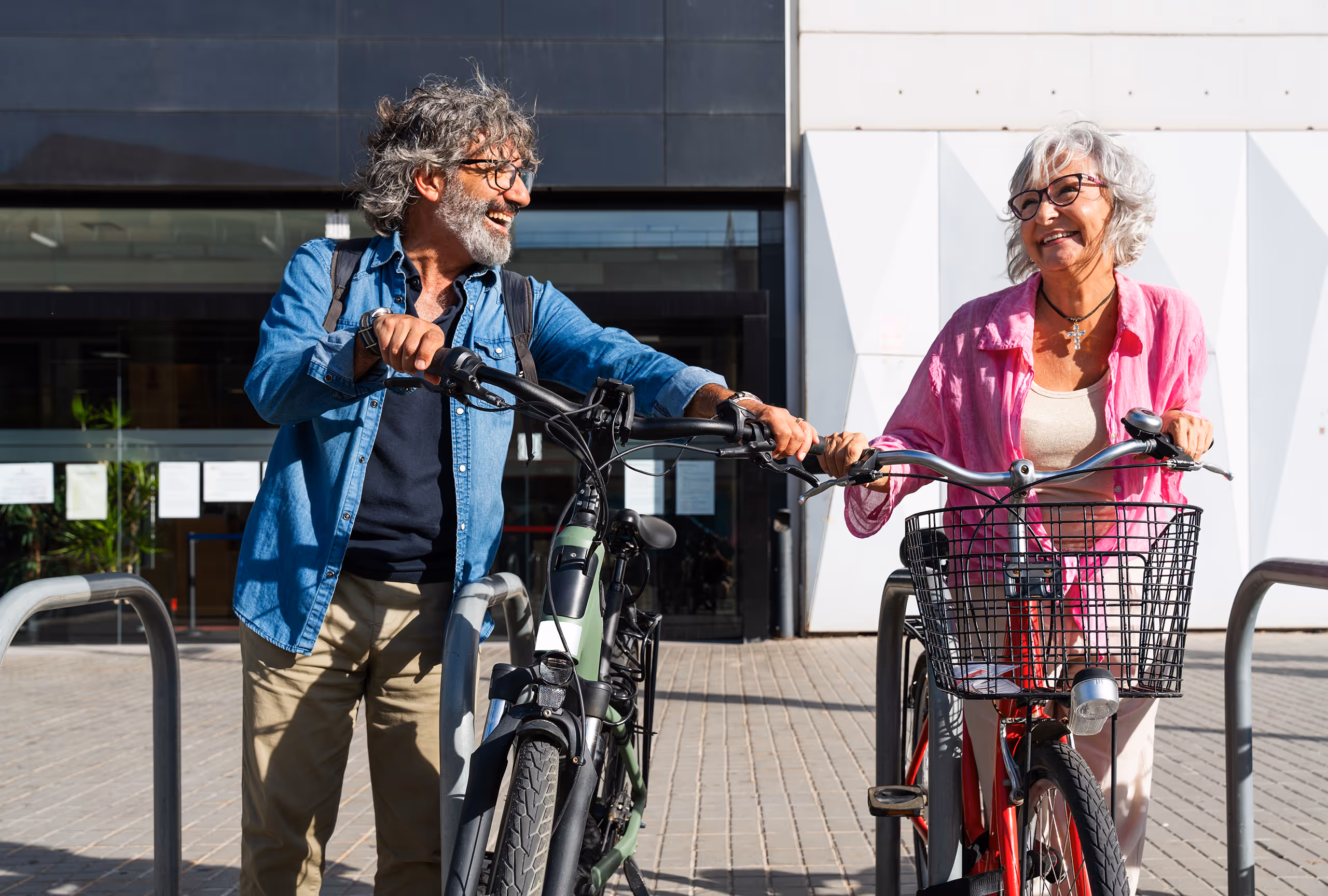 To smilende eldre mennesker står med hver sin sykkel. De ser glade på hverandre og nyter den solfylte dagen. Mannen har på seg en blå skjorte og kvinnen en rosa skjorte.