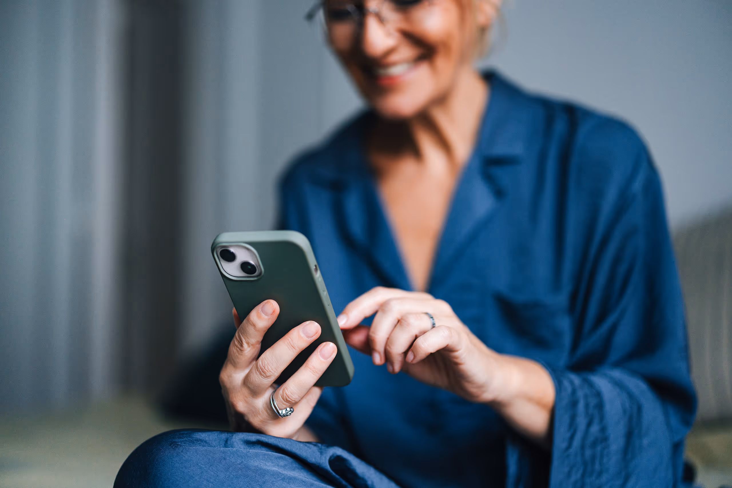 En smilende kvinne i blå pysjamas sitter og bruker en smarttelefon, mens hun fokuserer på skjermen. Bakgrunnen er uskarp, slik at oppmerksomheten rettes mot hånden hennes som interagerer med telefonen.