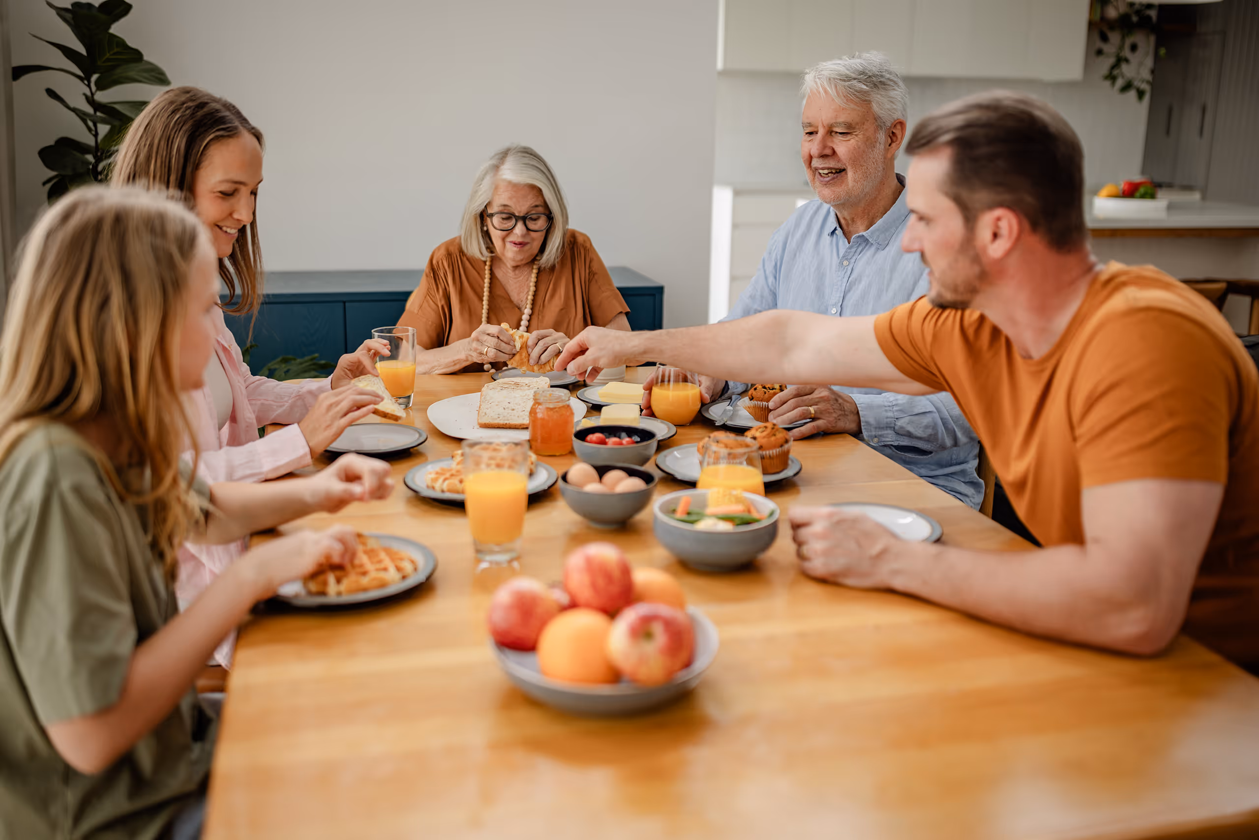 En familie på fem, inkludert to barn og tre voksne, sitter rundt et bord og spiser frokost sammen. Bordet er dekket med vafler, frukt, juice og annen mat, og alle ser glade og engasjerte ut.