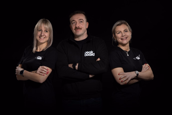 Three people in black peak board T-shirts stand side by side, smiling against a black background.