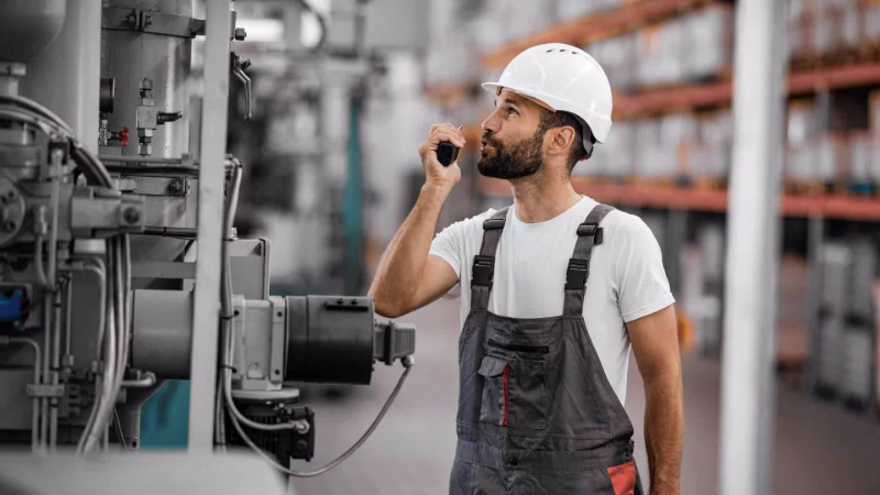 Techniker mit Schutzhelm nutzt ein Funkgerät vor einer Industrieanlage im Lager.