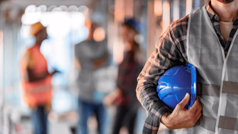 Construction worker with hard hat and high-visibility vest holds a safety helmet on a building site.