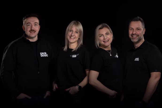 Vier Personen in schwarzen Peakboard-T-Shirts stehen nebeneinander und lächeln vor einem schwarzen Hintergrund.