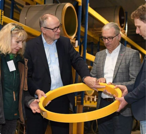 Geschäftsleute prüfen gemeinsam ein Bauteil in einer Produktionshalle zwischen Industrieanlagen.