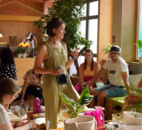 Frau erklärt Handarbeit in einem Workshop, Teilnehmende sitzen in gemütlicher Runde mit Bastelmaterialien.