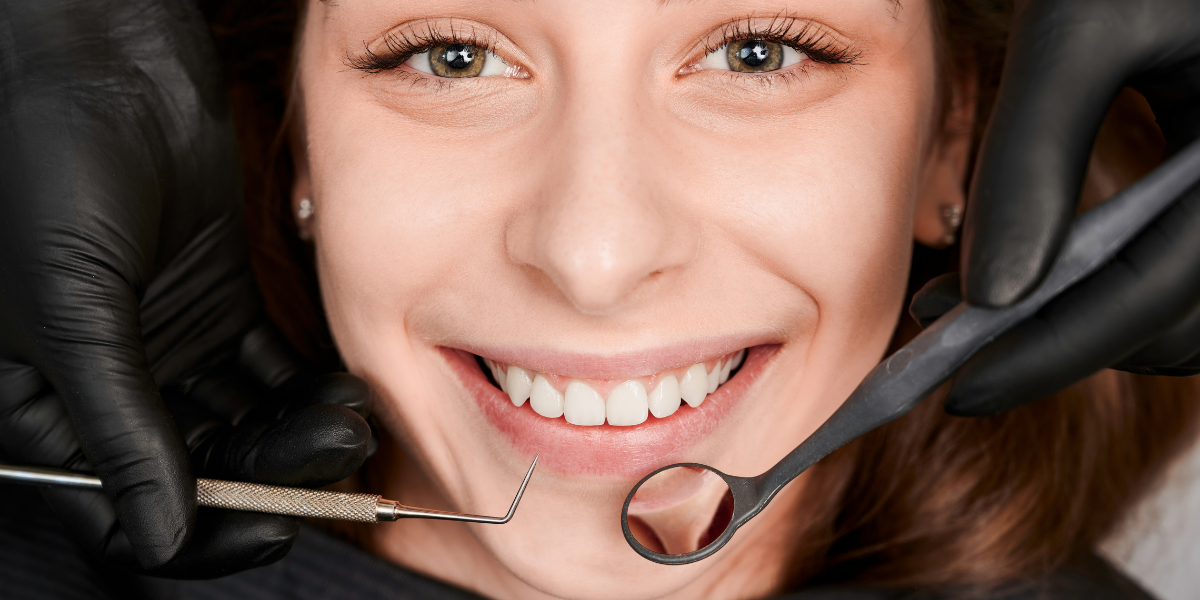 A smiling woman during a dental check-up, with a dentist wearing black gloves holding dental instruments near her teeth.