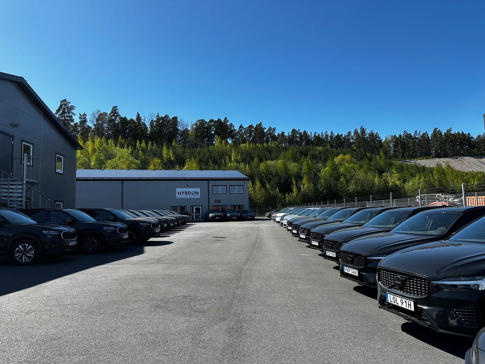 Flera svarta Volvo-bilar parkerade i två rader framför en byggnad med skylten HYRDON under en klarblå himmel.