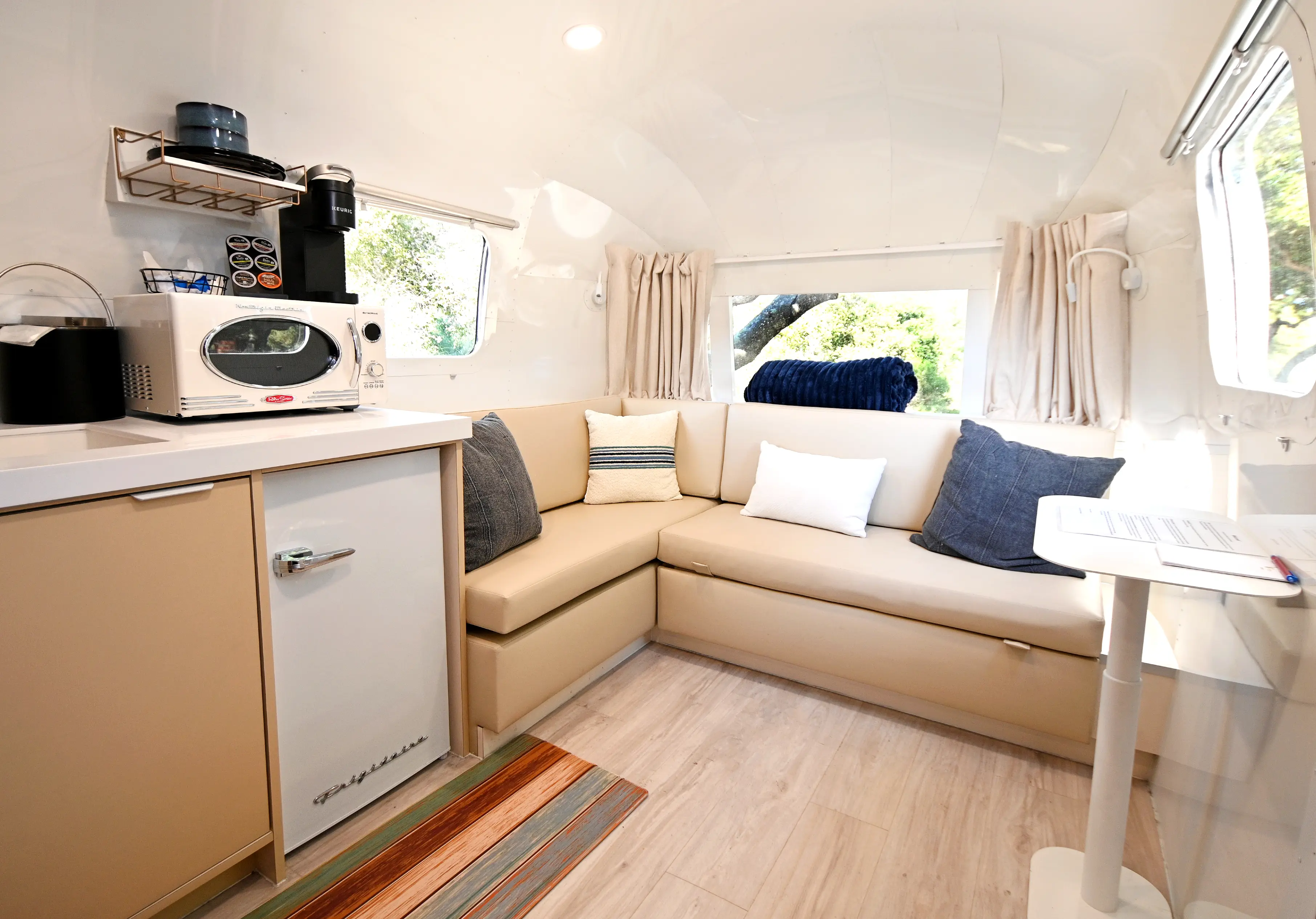 Cozy beige sectional sofa with pillows inside a bright camper next to a small kitchenette with a microwave.
