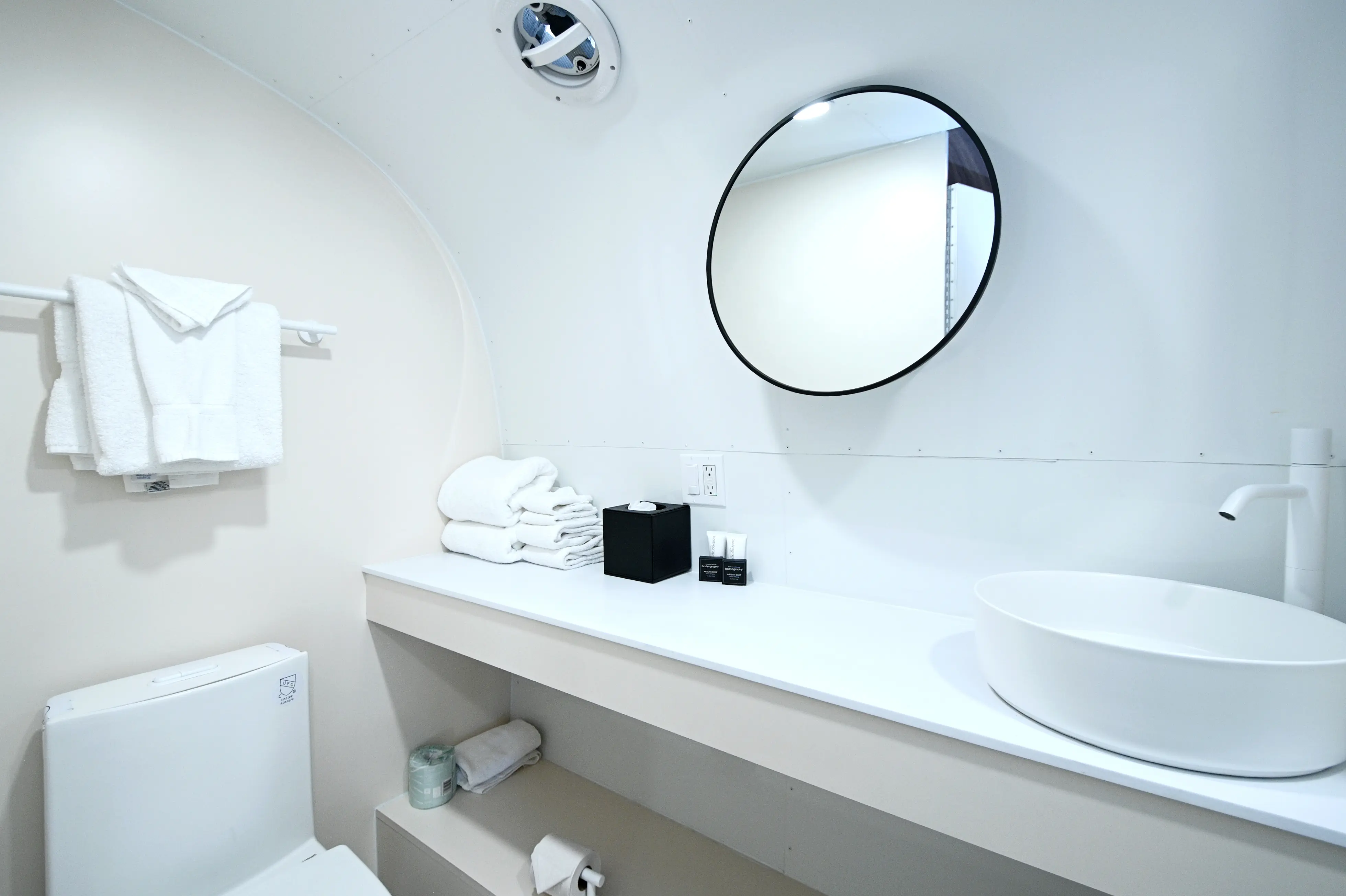 Minimalist white bathroom with round mirror, vessel sink, white towels, and toilet under towel rack.