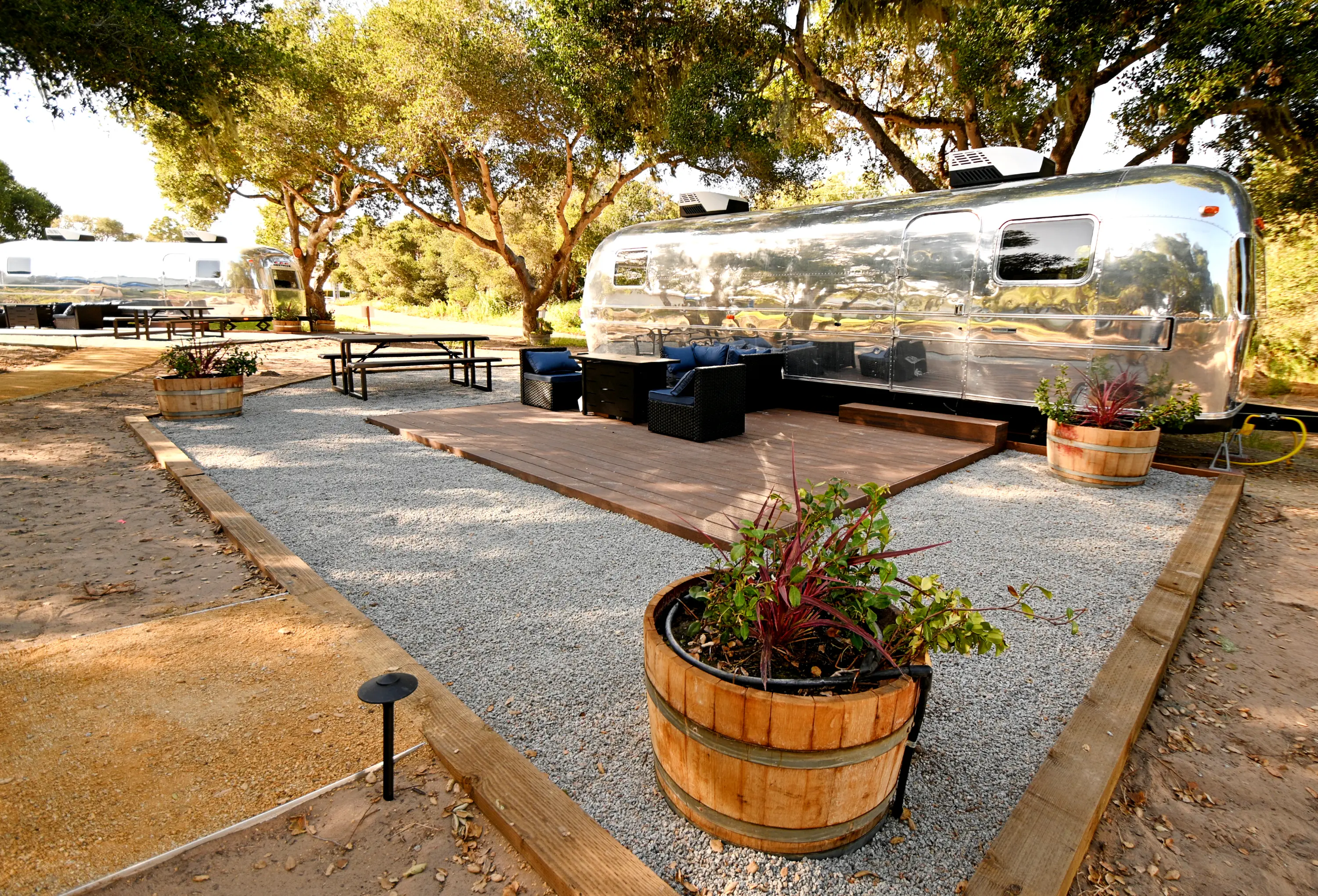 Outdoor seating area with wooden deck and metal chairs near shiny silver camper trailers under trees.