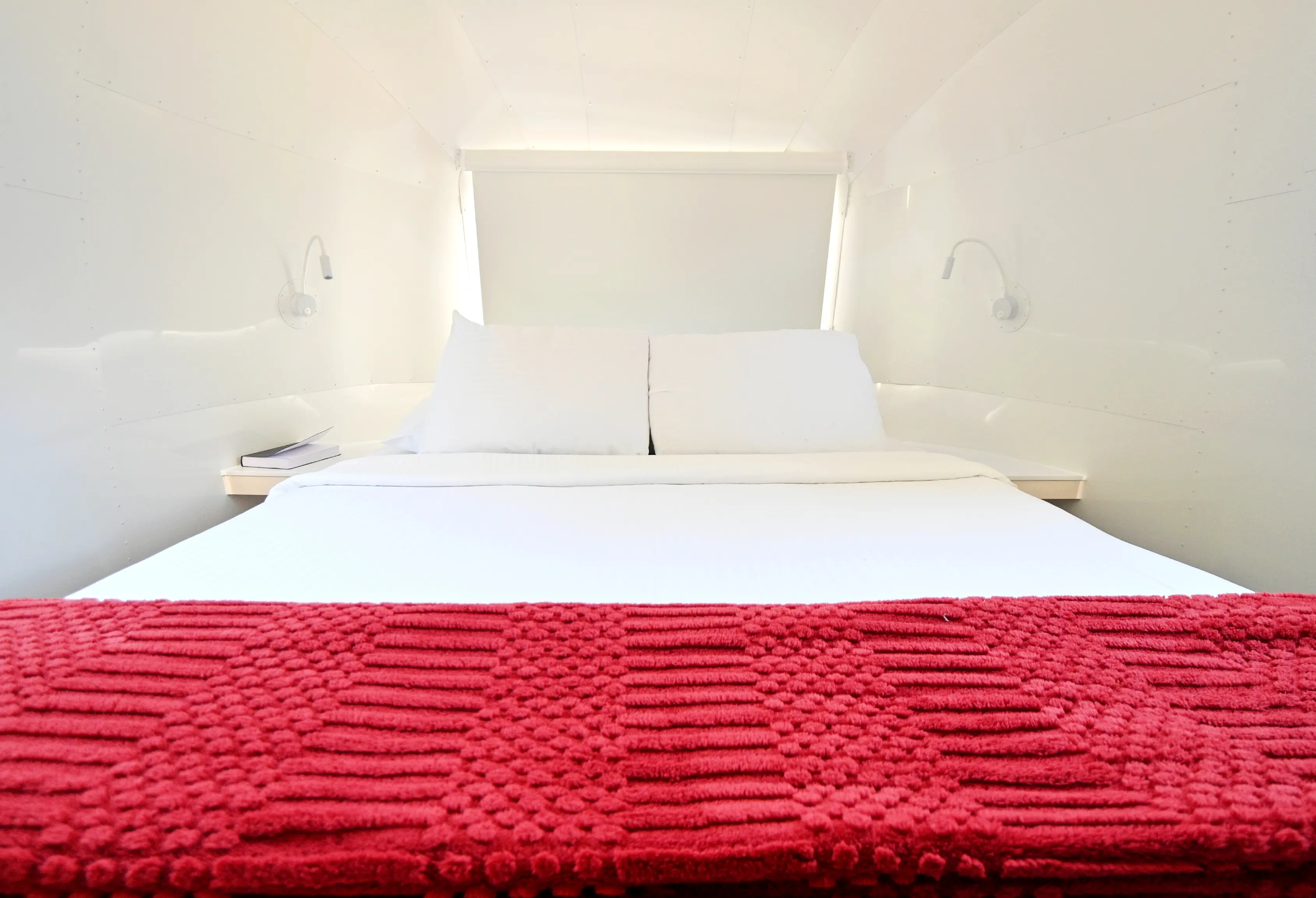 Minimalist white bed with two pillows, white bedding, a red textured blanket, and wall-mounted reading lights.