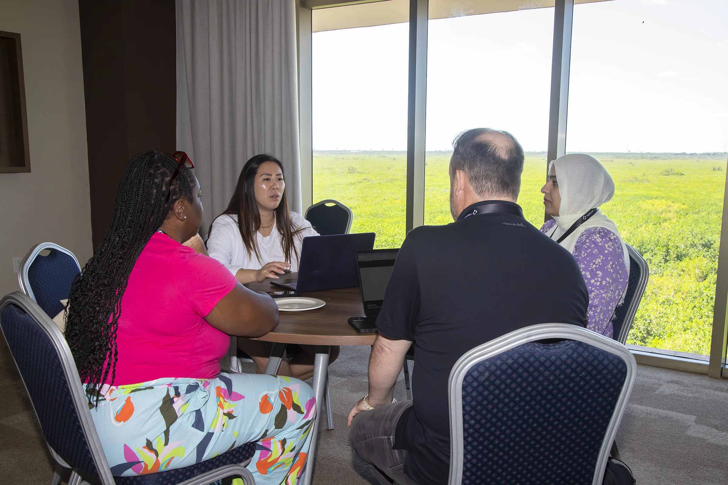 Four Replicant team members sit together at a round table with laptops, collaborating near a large window overlooking a bright green field.