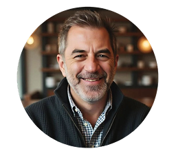 Smiling middle-aged man with gray hair and beard wearing a checkered shirt and dark sweater.