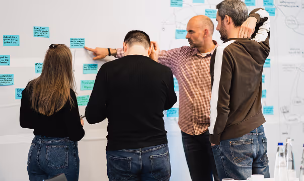 Fünf Personen stehen vor einer Wand mit Post-Its und diskutieren über ein Diagramm, das an der Wand hängt.