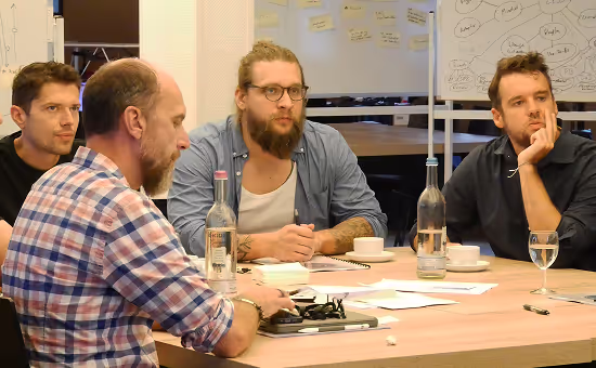 Vier Personen bei einer Besprechung, sitzen um einen Tisch und diskutieren intensiv, mit Notizen und Wasserflaschen auf dem Tisch.