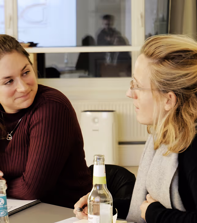 Zwei Frauen unterhalten sich bei einer Besprechung, beide lächeln und wirken in einem Gespräch vertieft, mit einem offenen Notizbuch auf dem Tisch.