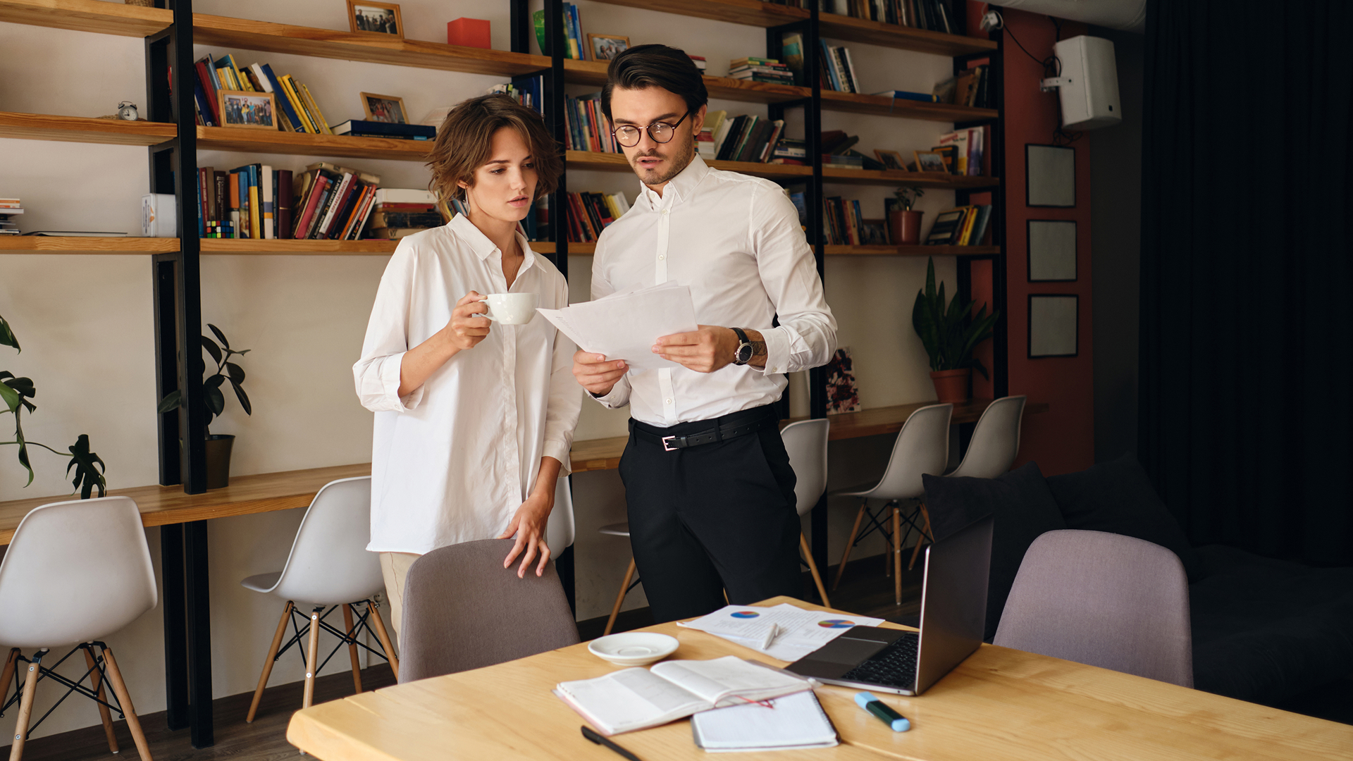 Two business colleagues standing in an office reviewing printed documents together, with a laptop and charts on the table in front of them