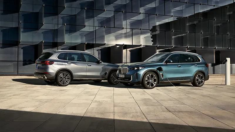 Zwei BMW X Modelle vor moderner Glasfassade, eines in Silber und eines in Petrol-Metallic an der Ladesäule – MOTORHEIMAT Wagner-Schwarz in Kirchseeon, Traunstein, Altenmarkt, Wasserburg und Oberhaching
