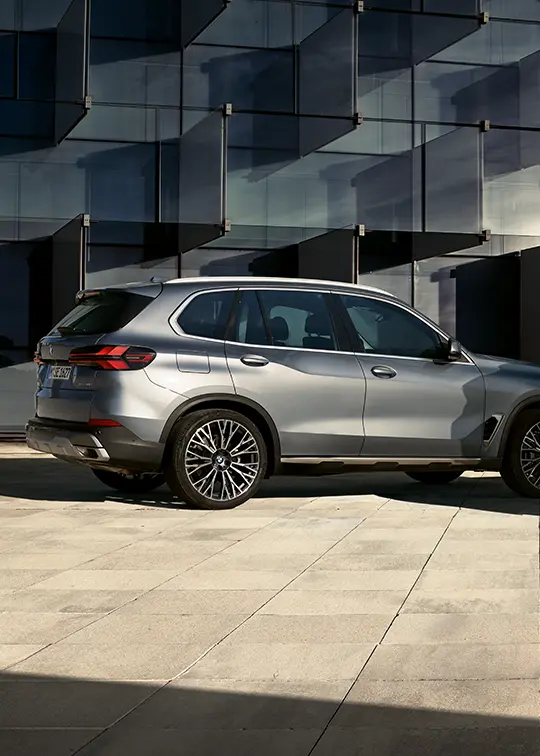 Zwei BMW X Modelle vor moderner Glasfassade, eines in Silber und eines in Petrol-Metallic an der Ladesäule – MOTORHEIMAT Wagner-Schwarz in Kirchseeon, Traunstein, Altenmarkt, Wasserburg und Oberhaching