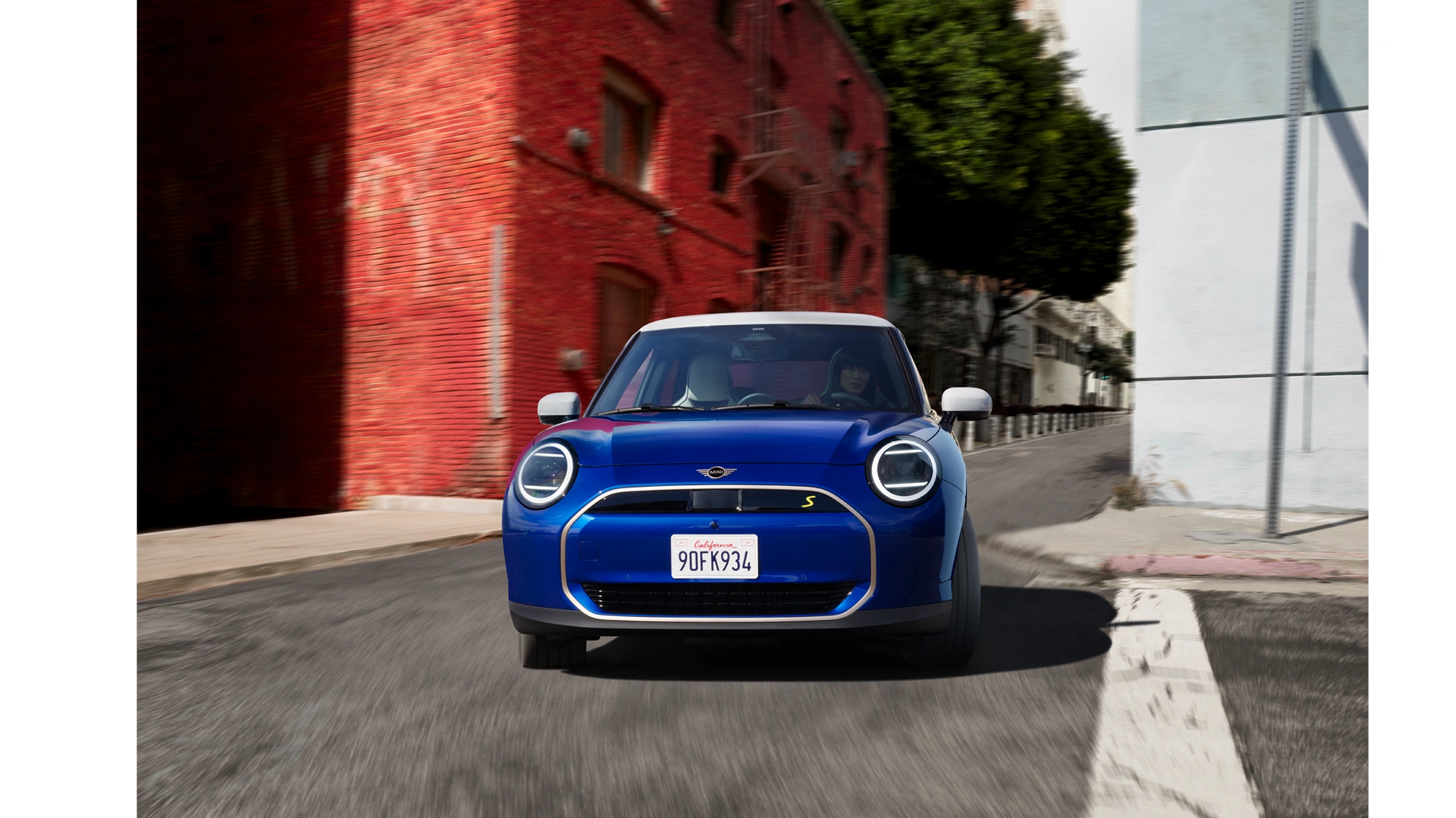 Frontalansicht eines blauen vollelektrischen MINI Cooper SE mit weißem Dach in Fahrt durch eine urbane Straßenschlucht mit roter Backsteinfassade – MOTORHEIMAT Wagner-Schwarz in Kirchseeon, Traunstein, Altenmarkt, Wasserburg und Oberhaching