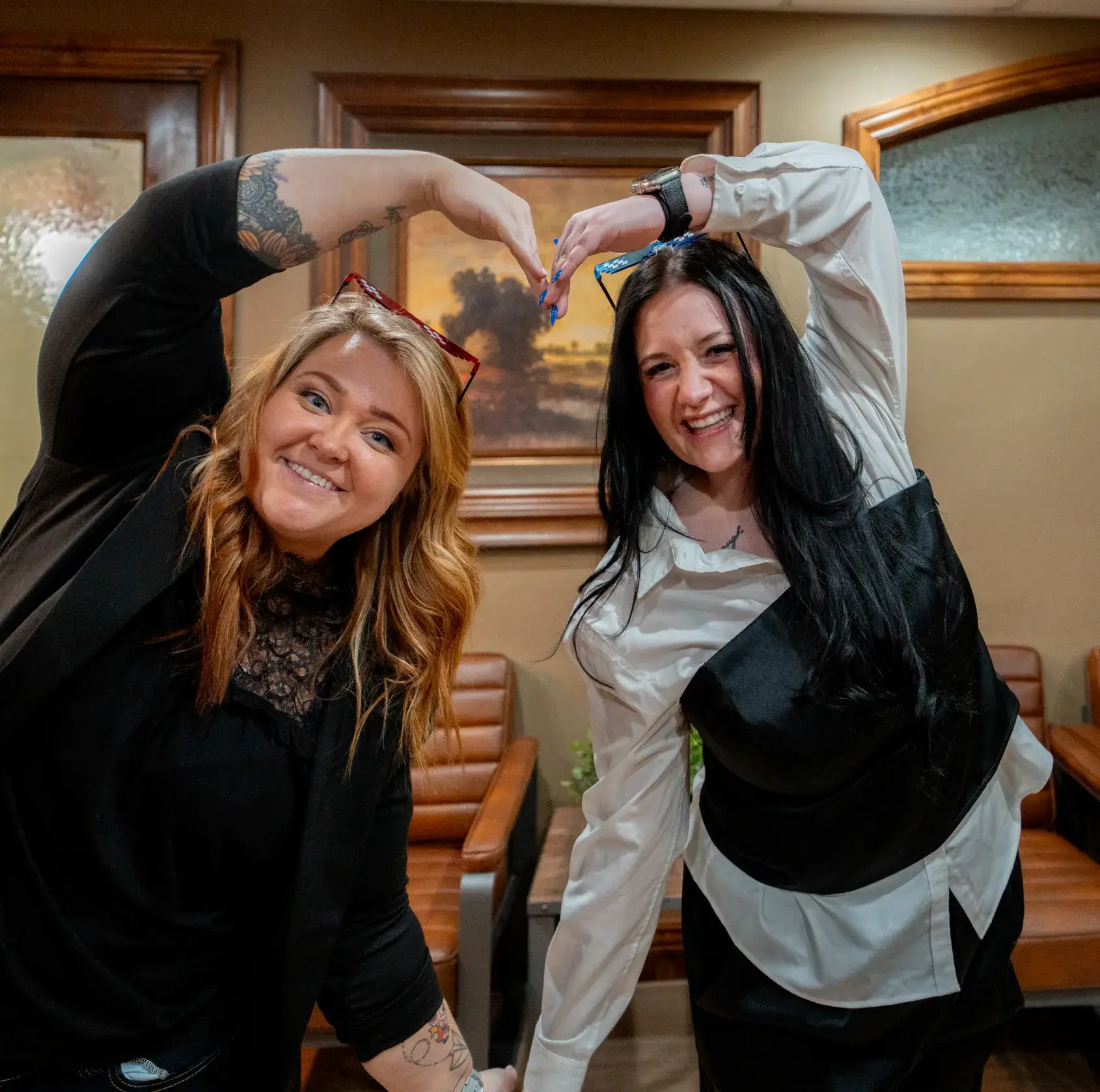 Two women smiling and forming a heart shape with their arms in an indoor setting.