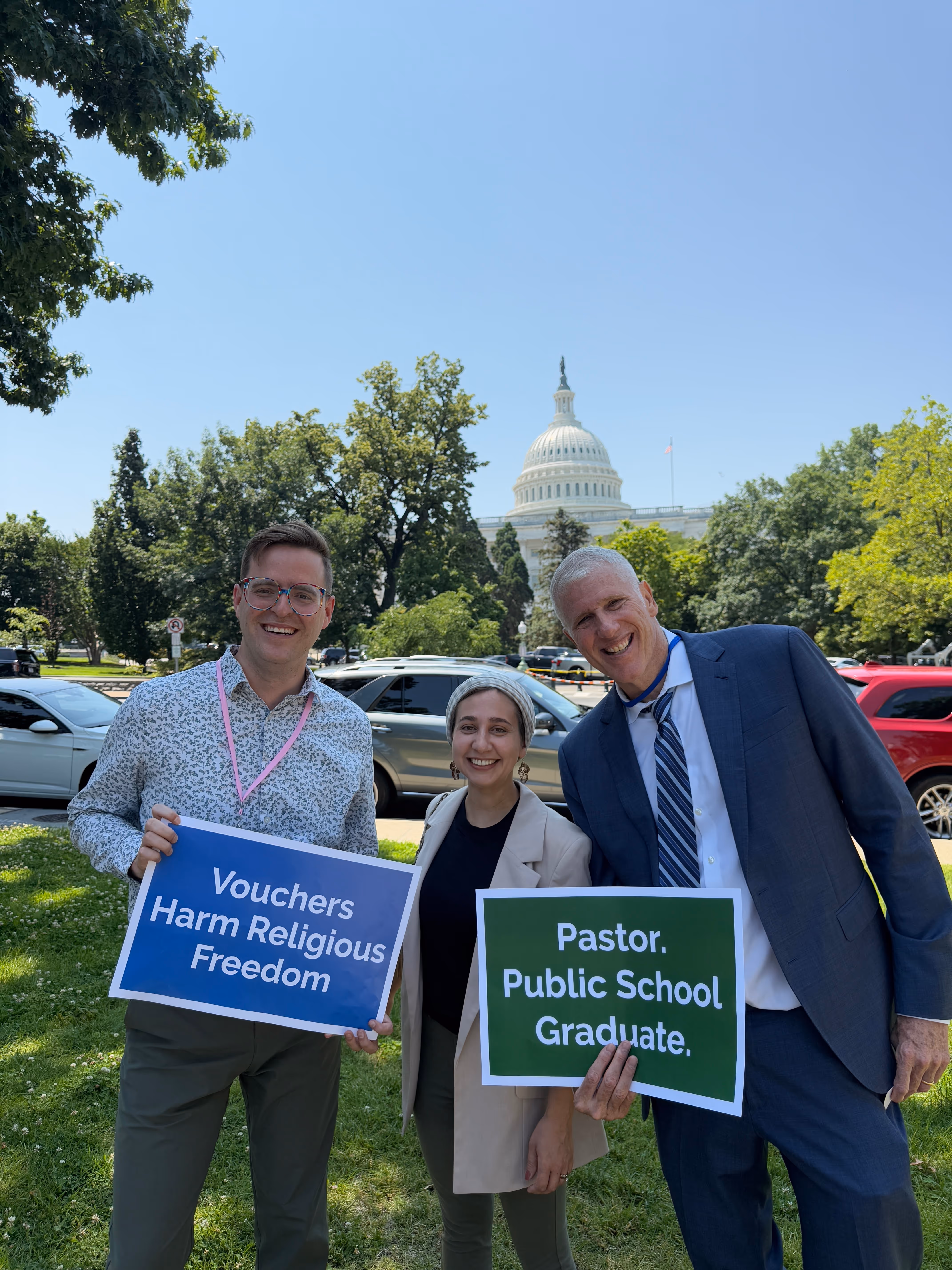 Group in front of the capitol holding signs: "Vochers harm religious freedom" and "Pastor. Public School. Graduate."
