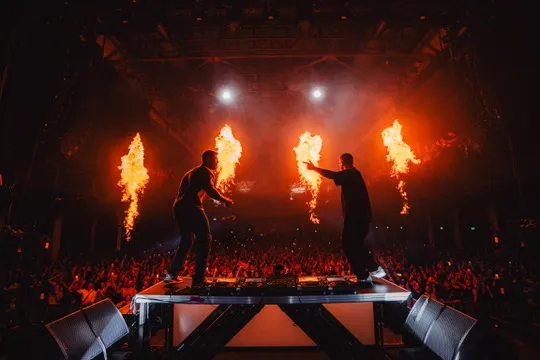 Zwei DJ Performen auf einem Podest mit einer Glasscheibe davor. im hintergrund sind flammen die vom Publikum bestaunt werden.