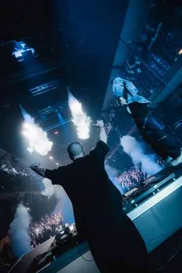 Ein DJ steht an seinem DJ Tisch, der mit Milchglas verkleidet ist. Über ihm ist ein Vorhang von blauen Licht zu erkennen, der in einem Quadrat angeordnet ist. Von oben ist Lichttechnik von photon zu sehen.