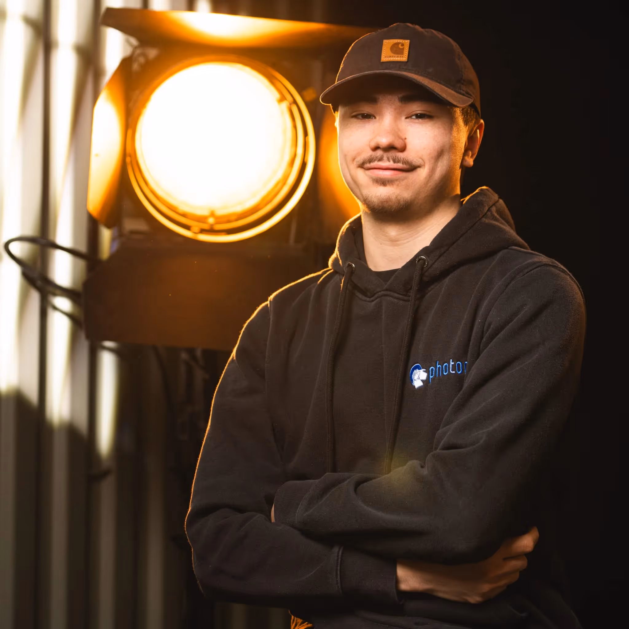 Fabian Blume mit schwarzen Haaren, trägt eine kappe und einem Schwarzen Hoody lächelt in die Kamera, eine Stufenlinse steht hinter ihm