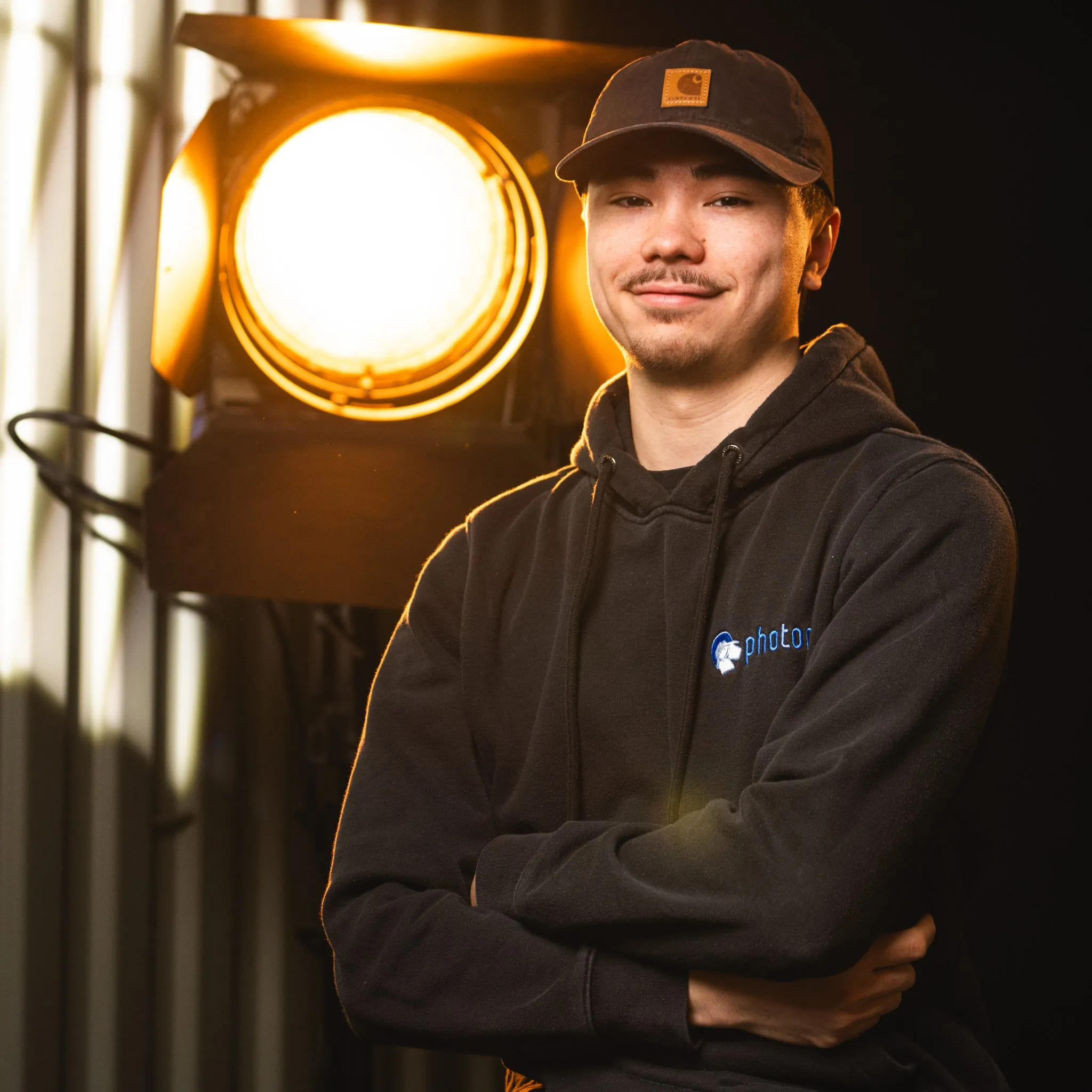 Fabian Blume mit schwarzen Haaren, trägt eine kappe und einem Schwarzen Hoody lächelt in die Kamera, eine Stufenlinse steht hinter ihm