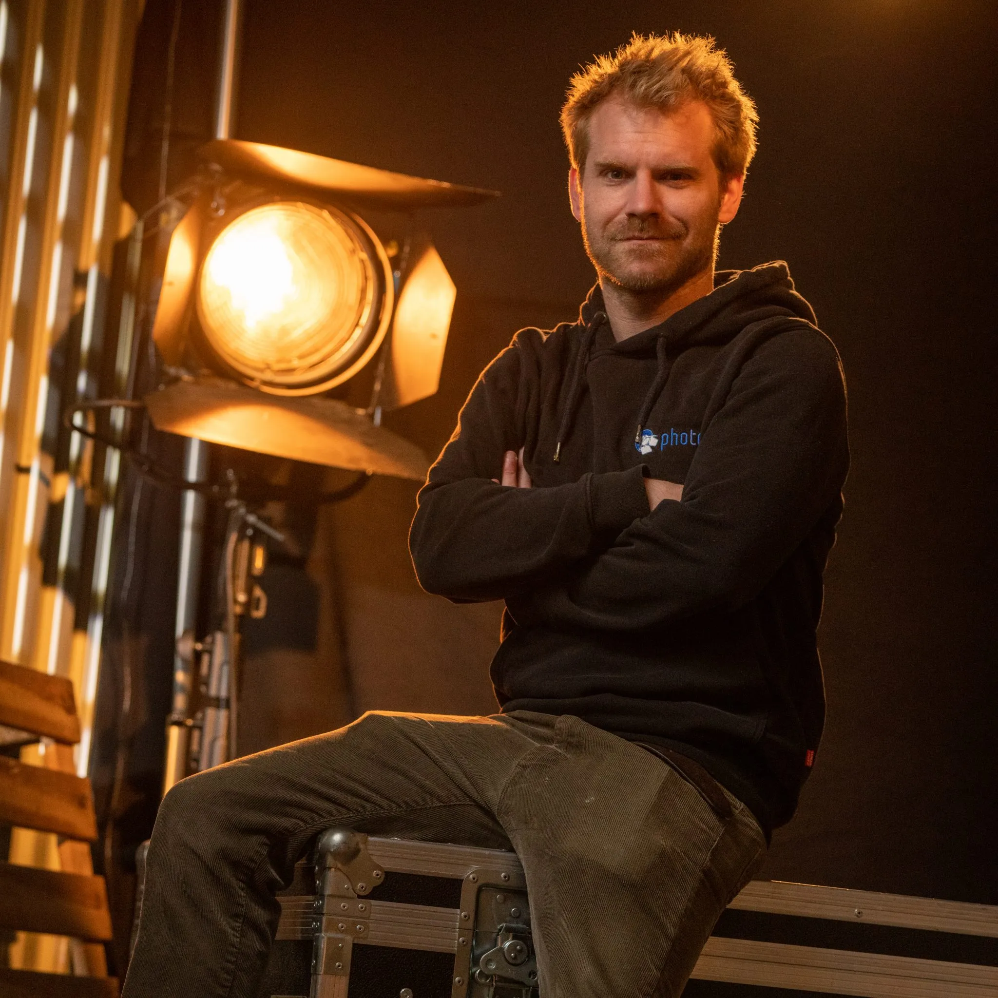 Christian Beyer mit blonden kurzen Haaren, trägt einen Schwarzen Hoody lächelt in die Kamera, eine Stufenlinse steht hinter ihm