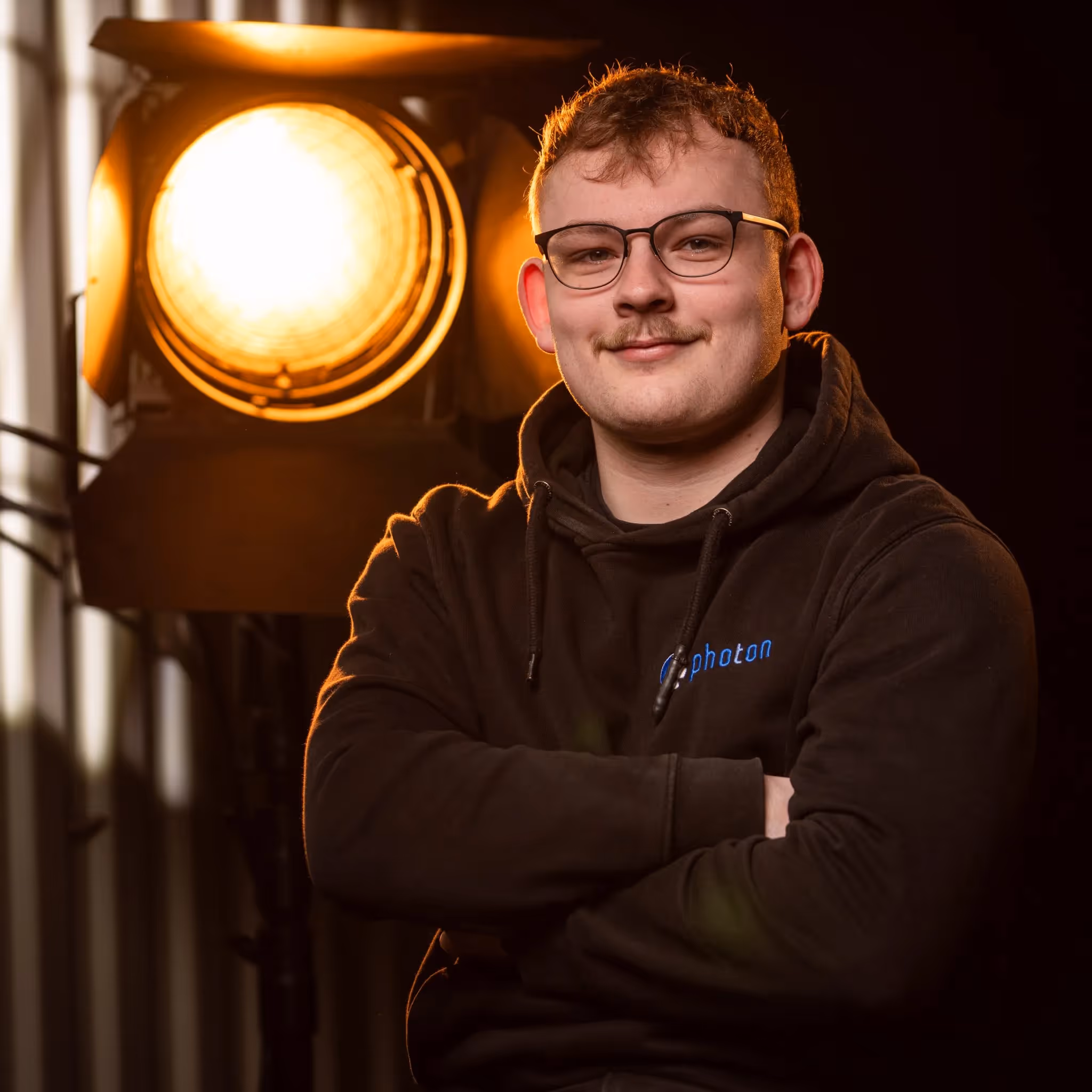Tim Kerzmann mit blonden Haaren und einem Schwarzen Pullover lächelt in die Kamera, eine Stufenlinse steht hinter ihm