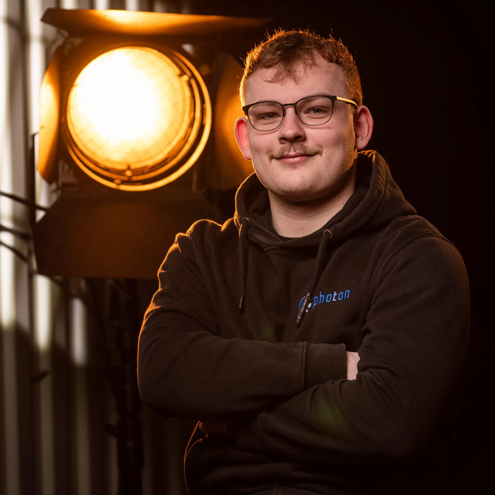 Tim Kerzmann mit blonden Haaren und einem Schwarzen Pullover lächelt in die Kamera, eine Stufenlinse steht hinter ihm