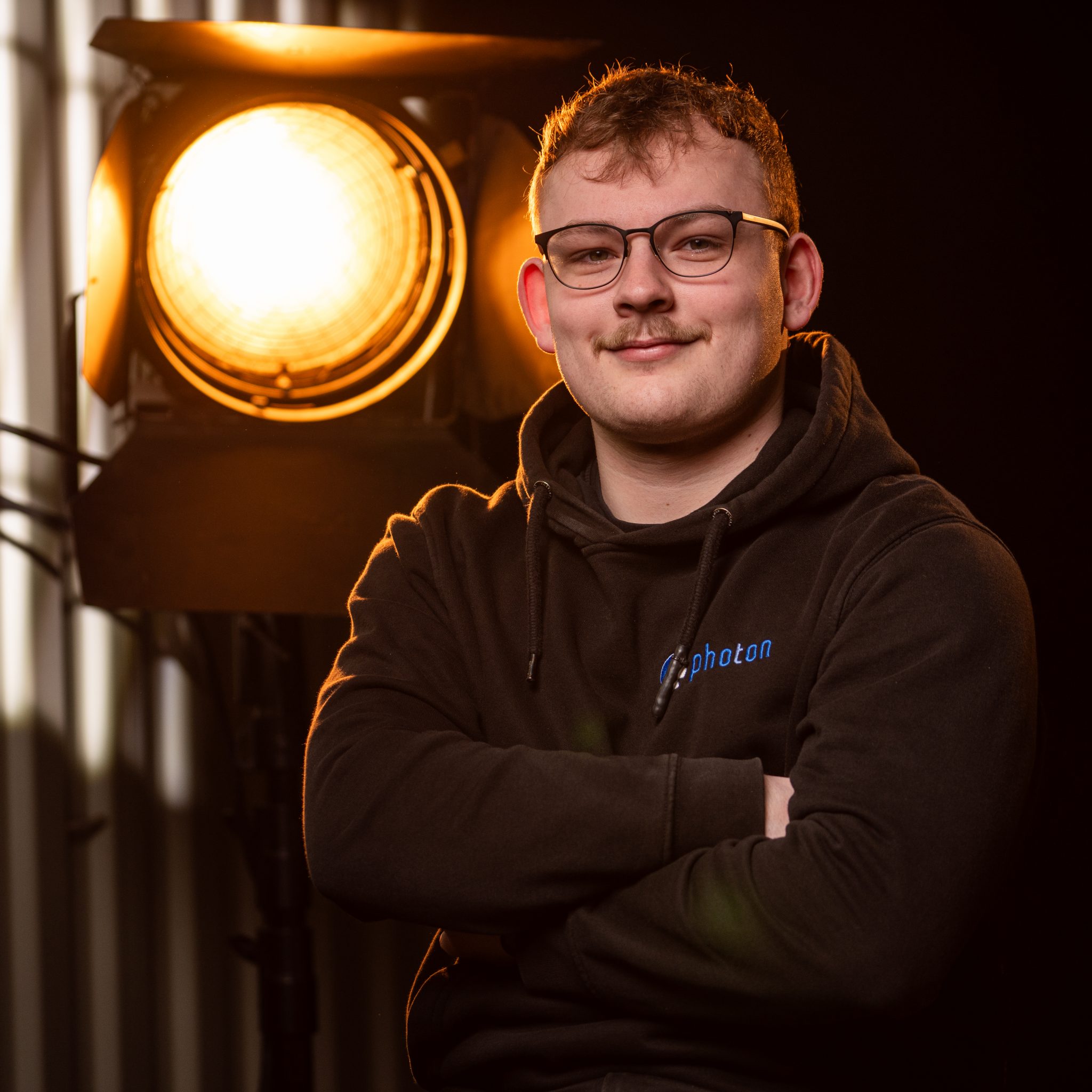 Tim Kerzmann mit blonden Haaren und einem Schwarzen Pullover lächelt in die Kamera, eine Stufenlinse steht hinter ihm