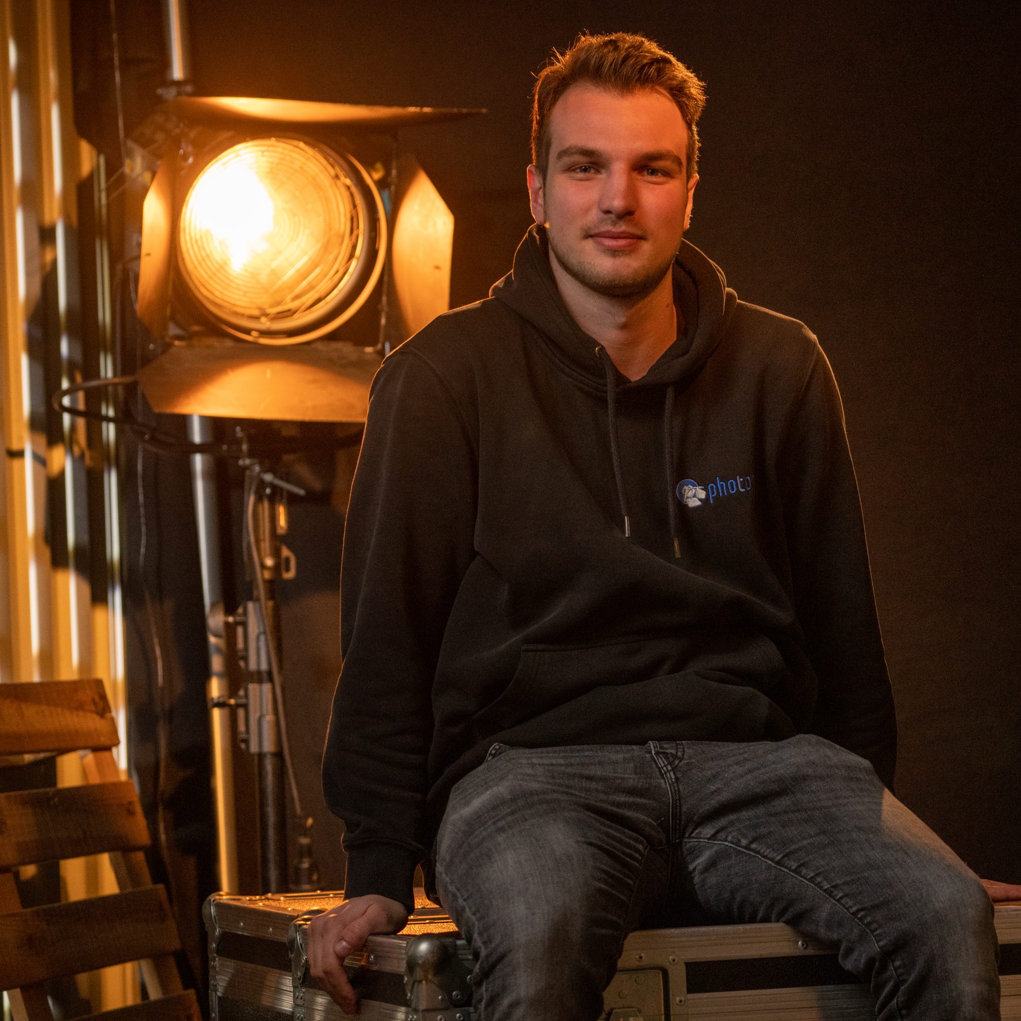 Nils Redlin mit schwarzen Haaren, trägt einen Schwarzen Hoody lächelt in die Kamera, eine Stufenlinse steht hinter ihm