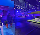 A blue-lit room with a glass booth front-left where a gamer streams, and a large blue-lit TV studio behind, by photon