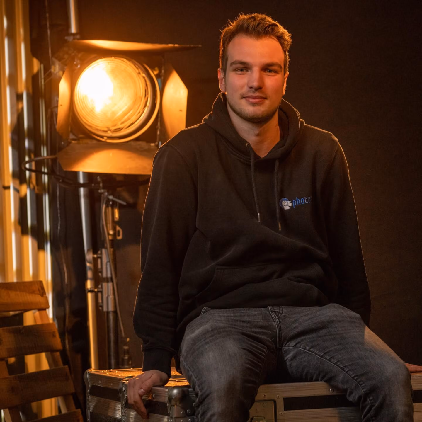 Nils Redlin mit schwarzen Haaren, trägt einen Schwarzen Hoody lächelt in die Kamera, eine Stufenlinse steht hinter ihm