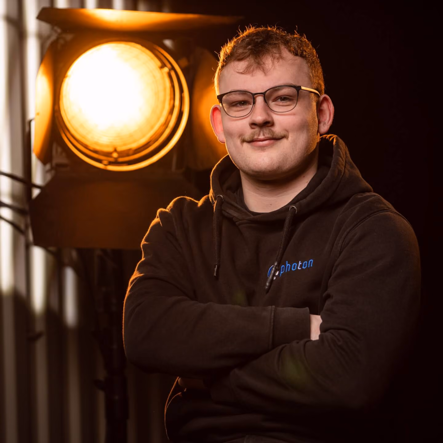 Tim Kerzmann mit blonden Haaren und einem Schwarzen Pullover lächelt in die Kamera, eine Stufenlinse steht hinter ihm