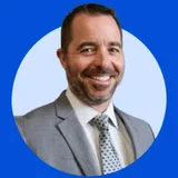 Smiling man with short dark hair and beard wearing a gray suit and dotted tie on a light blue circular background.