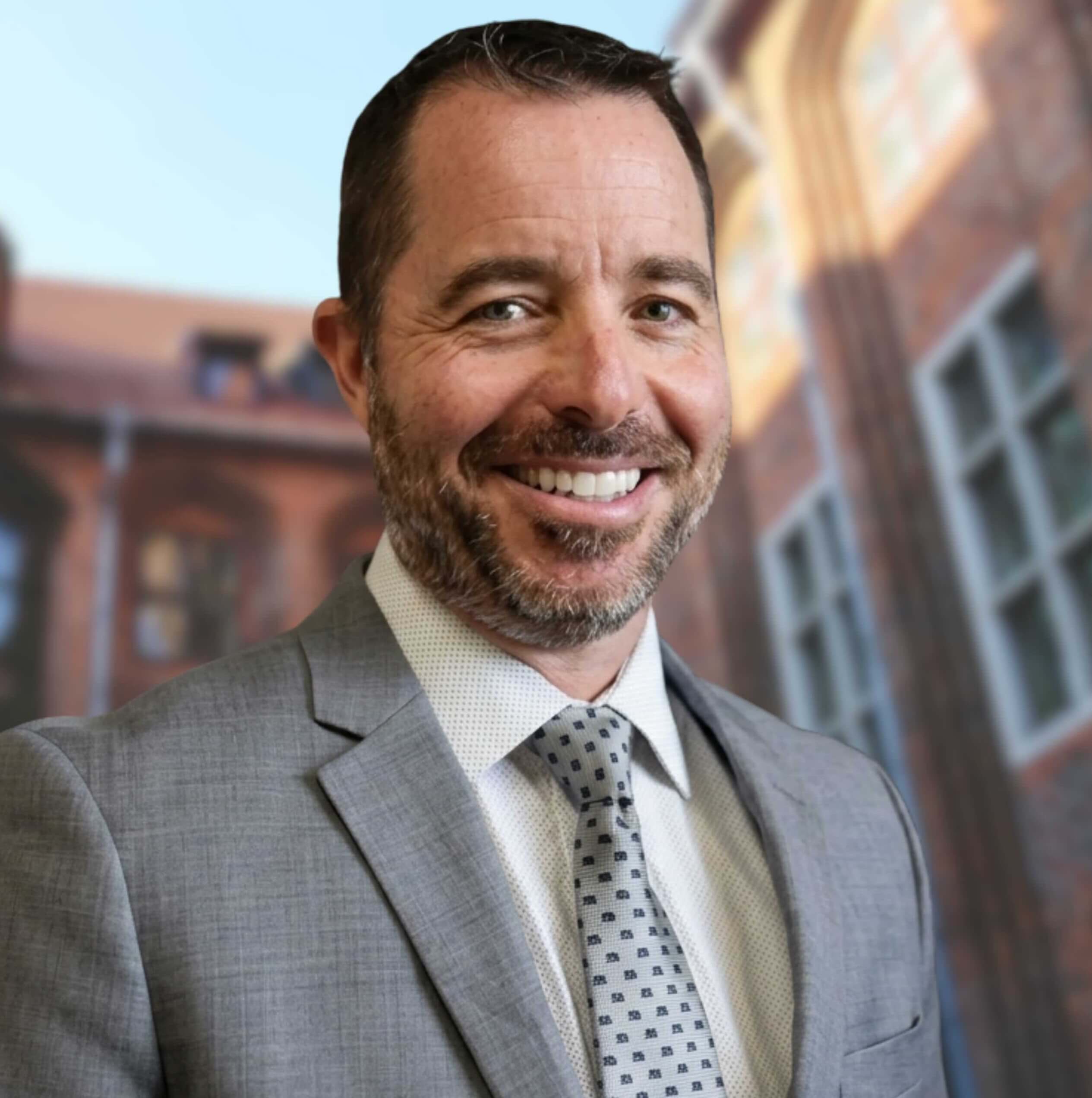 Smiling man with short hair and beard wearing a gray suit and patterned tie outdoors.