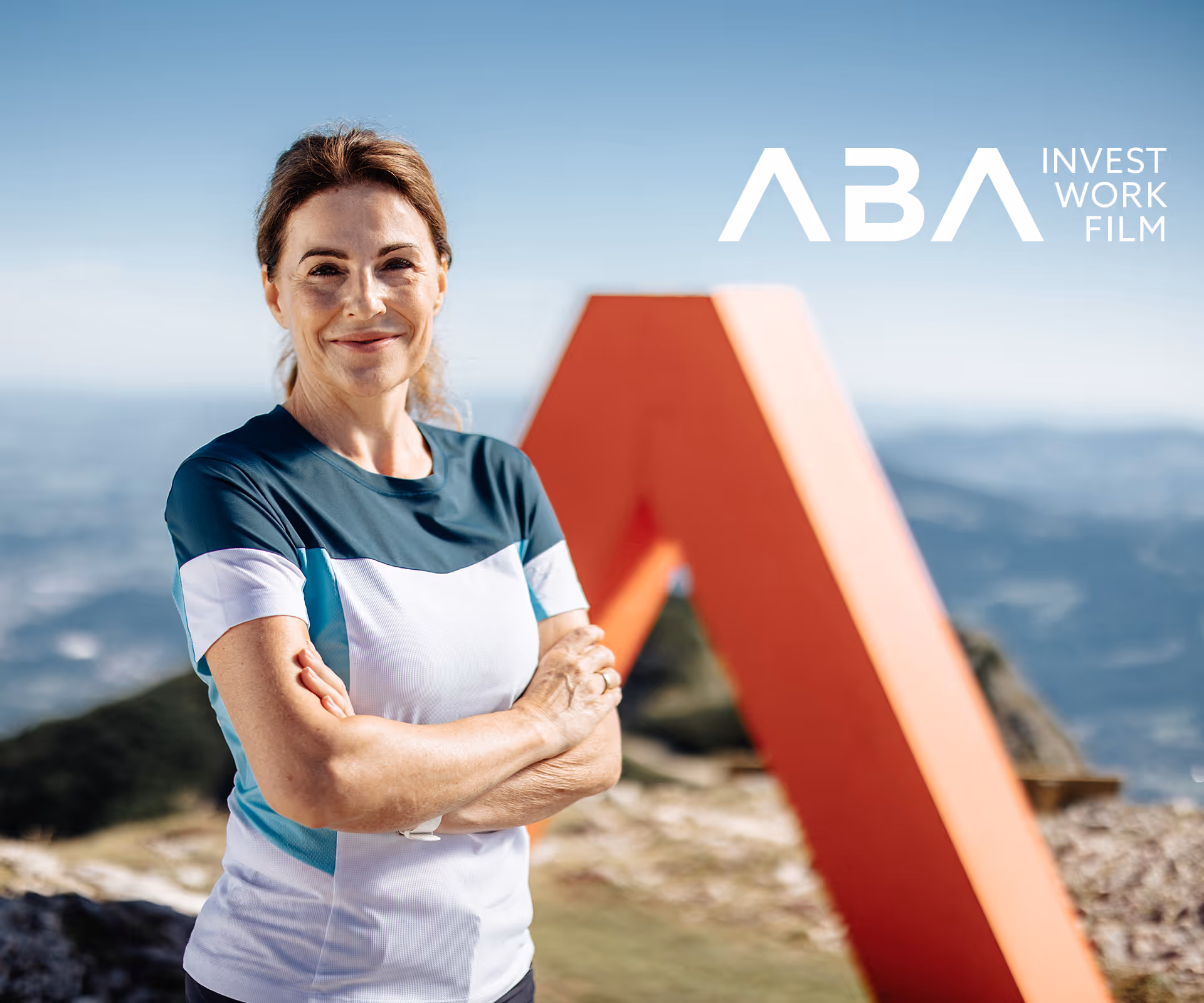 Frau in Sportkleidung vor rotem ABA-Signet am Berggipfel mit weitem Blick ins Tal. 