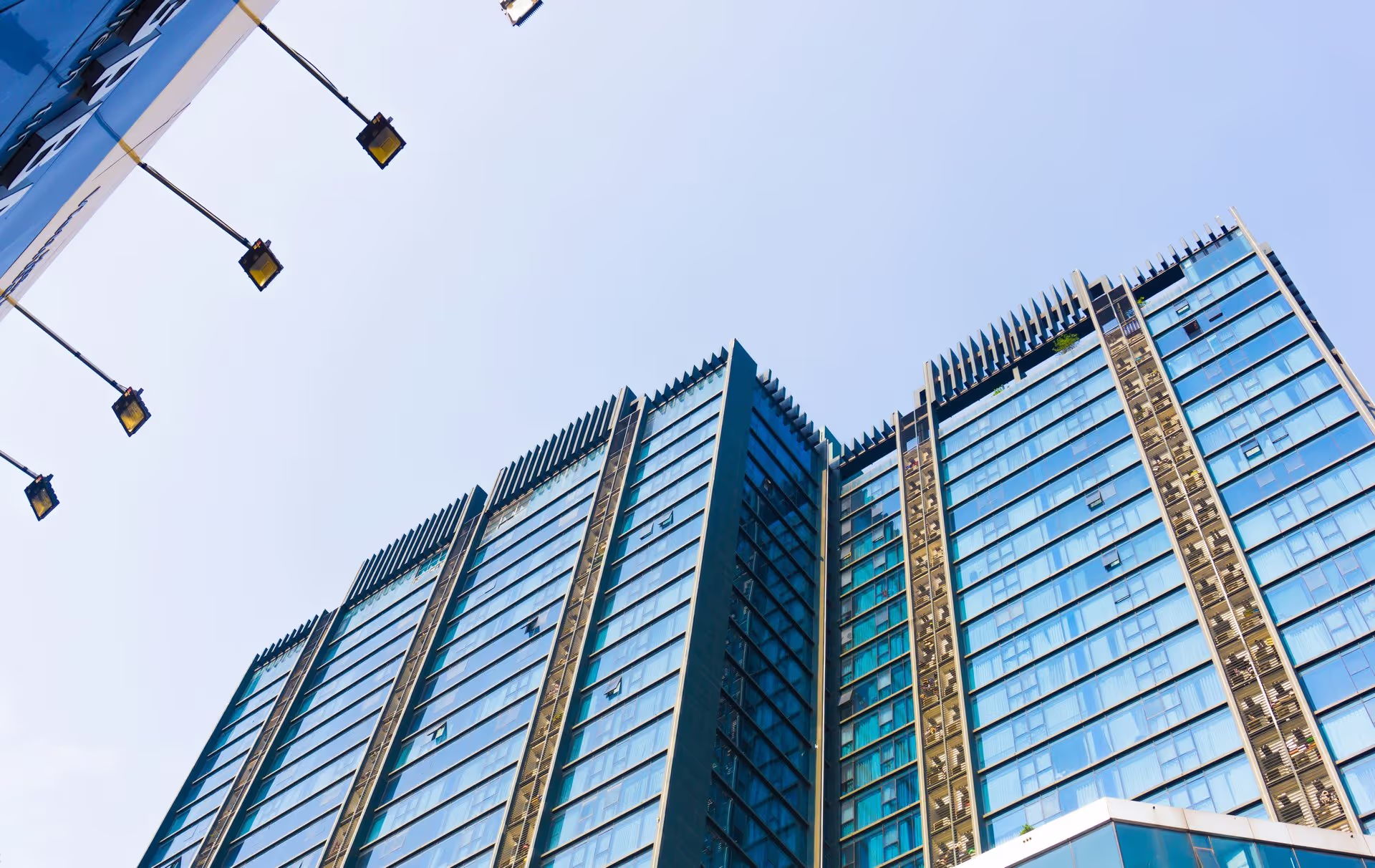 Tall modern office building with reflective glass windows against a clear blue sky, viewed from below. Streetlights visible on the left.