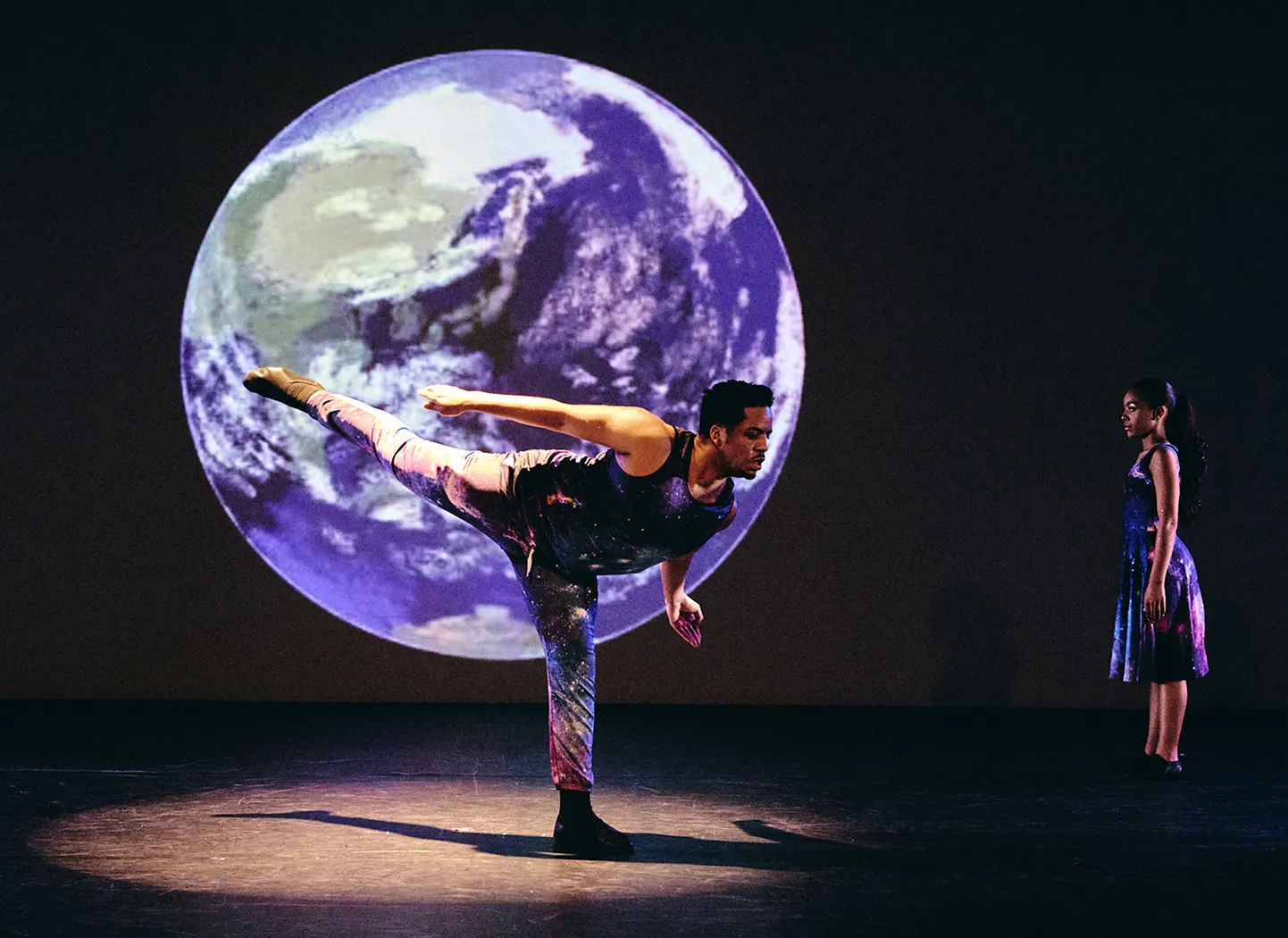 Two dancers on stage in galaxy-patterned costumes, one balancing on one leg with Earth projected in the background.