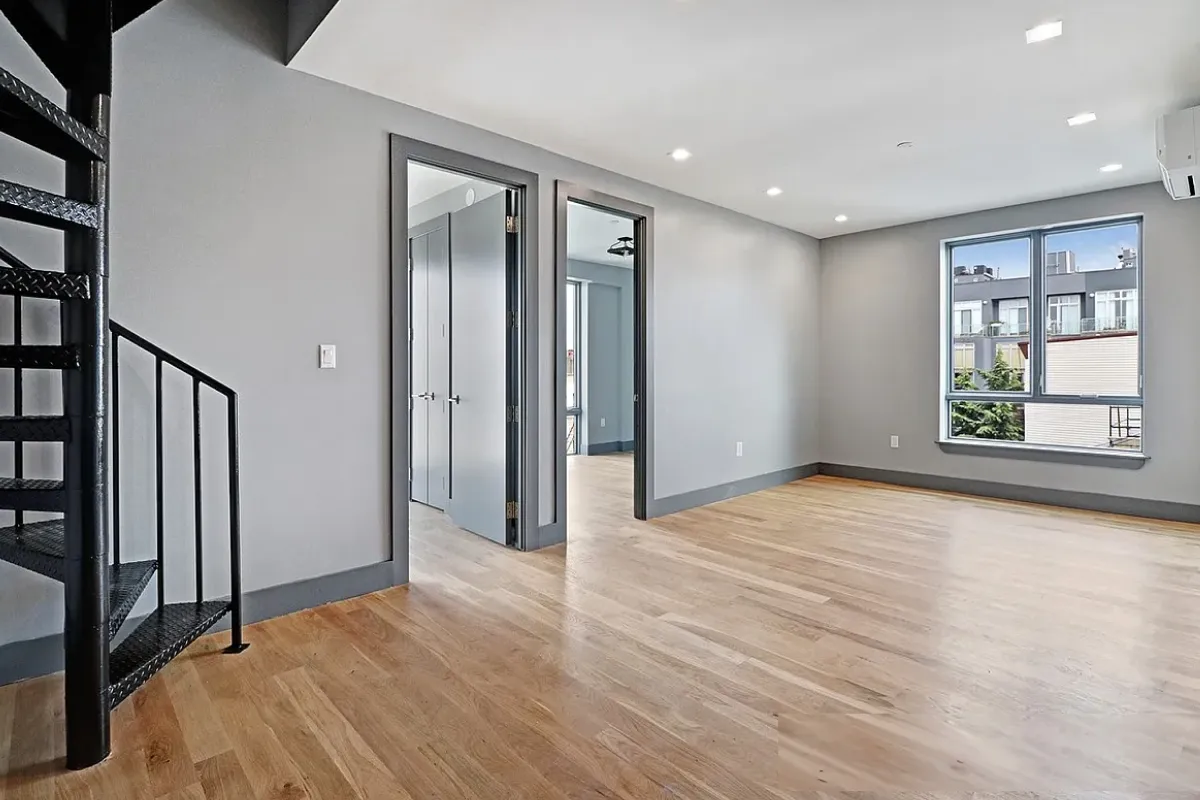 Empty modern room with light gray walls, wooden floor, spiral black metal staircase, two doorways, and a window showing adjacent buildings.