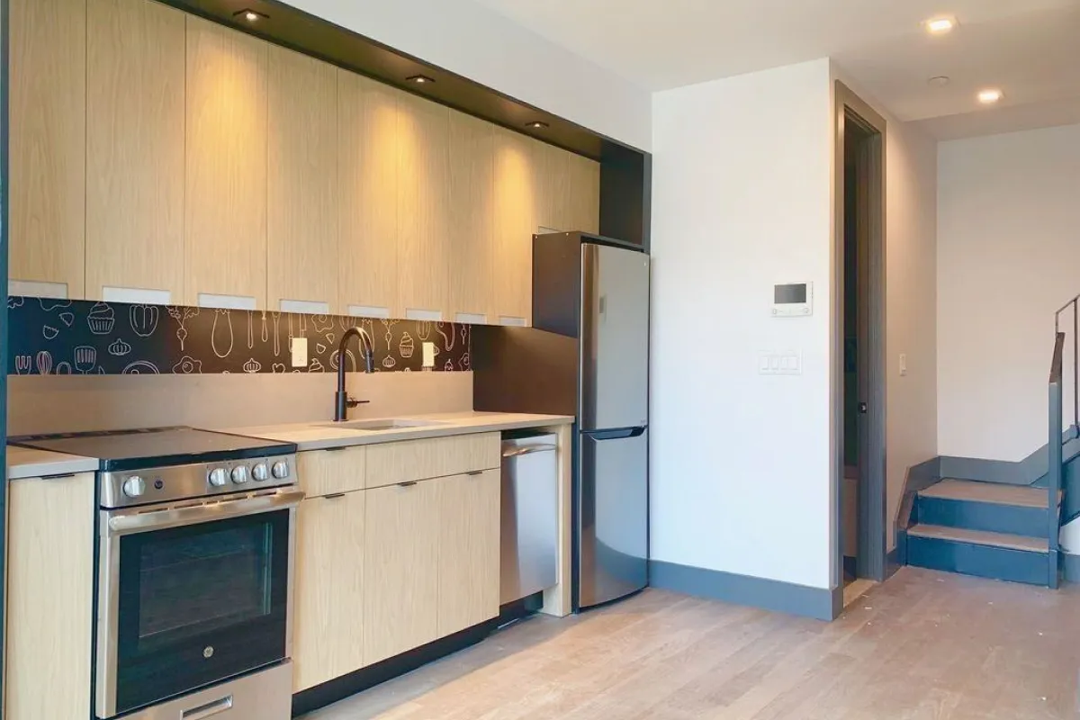 Modern kitchen with light wood cabinets, stainless steel oven, dishwasher, and refrigerator, black faucet, and decorative backsplash.
